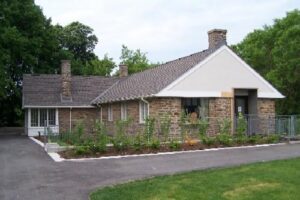 Image of the side of Canada’s Historic Places Riverwood, a historic stone building with a shingled roof, chimney, and landscaped garden, adjacent to a paved driveway.