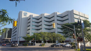 Image of the Whitby Courthouse, a modern multi-story building with a clean white facade, featuring the Ontario Court of Justice logo, surrounded by trees, parked cars, and traffic lights under a clear blue sky.