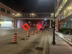 Nighttime safety zone beneath the pedestrian bridge at Garden City Tower, featuring orange cones, directional signs, and a lift, maintained by ICON Restoration.