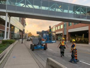 Daytime restoration crew at Garden City Tower pedestrian bridge, featuring workers in lifts and safety vests, with orange cones and a sunset backdrop, managed by ICON Restoration.
