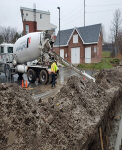 Image of a worker in a safety vest guiding a concrete mixer truck pouring into a foundation trench at a commercial site, with a brick building and traffic cones in the background.