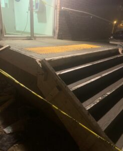 Image of a damaged staircase under repair at night, cordoned off with yellow tape, outside a brick building with glass doors, showcasing ICON Restoration's emergency work.