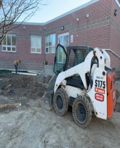 Image of a white skid steer loader (S175 model) working on a construction site in front of a brick building, with a worker handling materials under a clear sky.