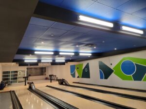 A modern bowling alley with polished wooden lanes, a teal ceiling with energy-efficient LED lights, and colorful abstract murals on the walls, enhanced by ICON Restoration at Maple Community Centre.