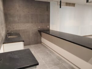A modern kitchen at Maple Community Centre with black granite countertops, a stainless steel sink, and gray tiled walls and floor, showcasing the recent renovation by ICON Restoration.