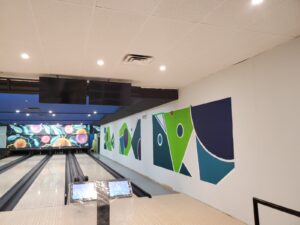 A modern bowling alley with polished wooden lanes, colorful abstract murals in green, blue, and purple on the walls, overhead screens, and energy-efficient lighting, upgraded by ICON Restoration at Maple Community Centre.