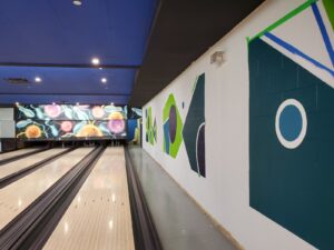 A modern bowling alley featuring polished lanes, a colorful mural with abstract shapes and glowing designs, and a teal ceiling with energy-efficient lighting, revitalized by ICON Restoration at Maple Community Centre.