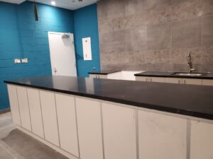 A modern kitchen with a sleek black countertop, white cabinets, and a teal brick wall, featuring a sink, tiled backsplash, and energy-efficient lighting, reflecting the recent restoration by ICON Restoration at Maple Community Centre.