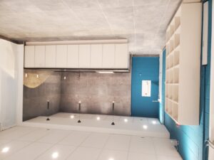 A modern kitchen with white cabinetry, gray tiled walls, a teal door, and recessed lighting with pendant fixtures, renovated by ICON Restoration at Maple Community Centre.