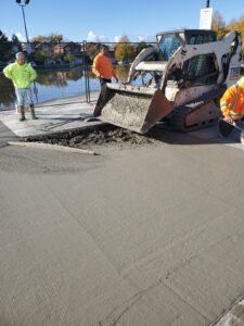 Image of the Chancellor Community Centre restoration project in Vaughan by ICON Restoration, featuring workers in safety gear pouring concrete with a backhoe near a newly laid sidewalk, surrounded by a fenced area, water, and residential buildings under a clear sky.