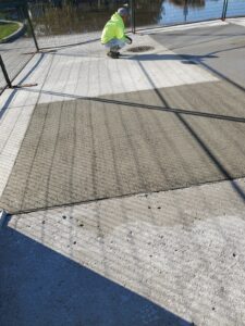 Image of a worker in a high-visibility yellow jacket inspecting freshly poured concrete at the Chancellor Community Centre restoration site in Vaughan, with a fenced area, water, and shadows cast on the new sidewalk under a clear sky.