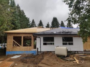 Image of a residential renovation by ICON Restoration, showing a home with a partially constructed wooden extension, new roof, and large window openings, surrounded by a pile of dirt and trees under a cloudy sky.