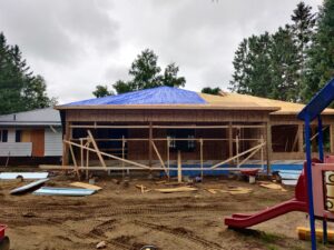 Image of a residential extension under construction by ICON Restoration, featuring a wooden-framed structure with a blue tarp roof, large windows, and scattered construction materials, with a playground slide visible in the background.