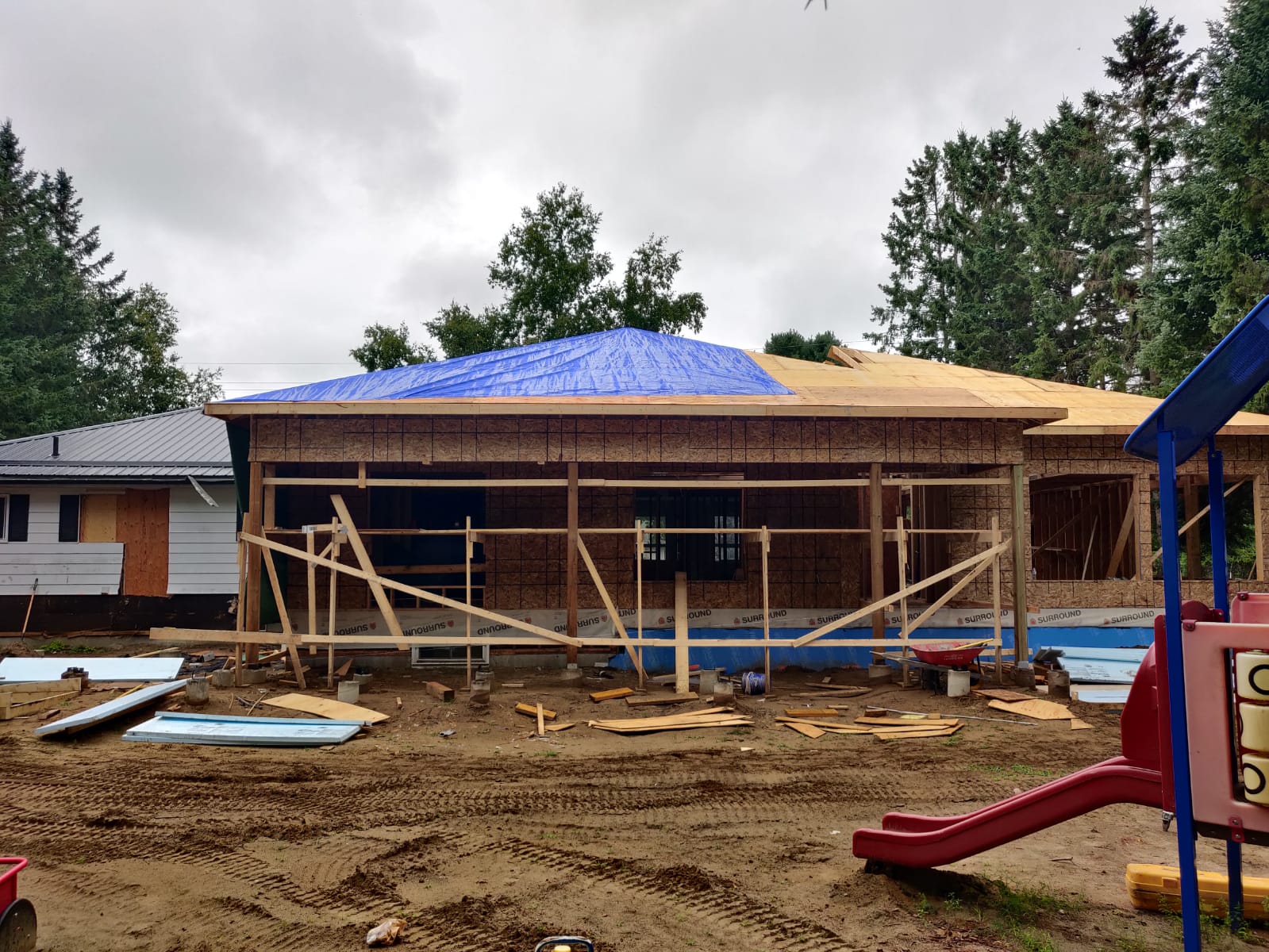 Image of a residential extension under construction by ICON Restoration, featuring a wooden-framed structure with a blue tarp roof, large windows, and scattered construction materials, with a playground slide visible in the background.