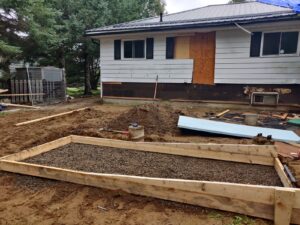 Image of an outdoor renovation by ICON Restoration, featuring a residential backyard with a wooden-framed foundation filled with gravel, a house with boarded windows, and scattered construction materials under a cloudy sky.