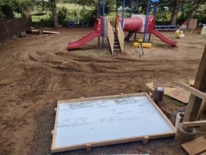 An outdoor play area at RAA-1815 Minden Daycare Centre with a partially constructed foundation, a colorful playground with red slides and blue frames, and construction equipment, renovated by ICON Restoration.