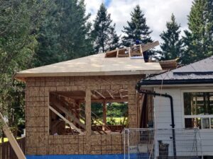 Image of a residential addition under construction by ICON Restoration, showing a wooden-framed extension with exposed beams, a partially completed roof, and large window openings, set against a backdrop of trees and an existing home.