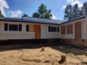 Image of a residential home exterior under renovation by ICON Restoration, showing a white house with a dark metal roof, boarded-up windows, exposed wooden framing, and a sandy yard with concrete blocks, highlighting ongoing modernization and structural improvements.