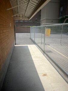 Covered concrete walkway next to a brick building, leading to a fenced-off construction area with a yellow sign at 2 Surrey Place.