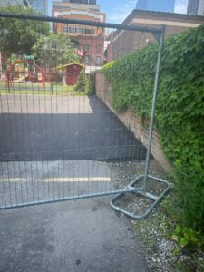 Fenced-off paved area at 2 Surrey Place with a playground and modern skyscrapers in the background next to a brick building.