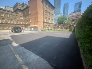 Newly paved area at 2 Surrey Place with a playground featuring red equipment, a parked vehicle, and modern skyscrapers in the background next to a brick building.