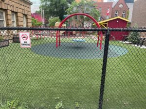 Fenced playground at 2 Surrey Place with artificial turf, red arch, blue swing, and a 'No Smoking' sign near a brick building.
