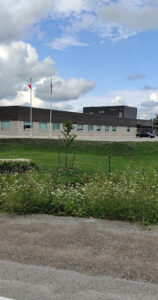 Image of an Ontario Provincial Police facility with upgraded grounds, featuring a modern building, well-maintained green areas, and two flagpoles under a partly cloudy sky.