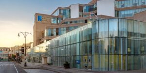 Image of the Hamilton Public Library, a modern building with a prominent glass facade, featuring the library logo, large windows, and a street view at sunset with streetlights and urban surroundings.