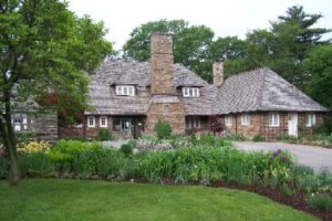 Image of Canada’s Historic Places Riverwood, a historic stone building with shingled roofs, multiple chimneys, and well-maintained gardens, surrounded by lush greenery and a paved driveway.