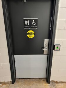 Accessible restroom door at Niagara College Canada’s Welland Campus, with an automatic door sign, restroom and wheelchair symbols, and a green activation button, post-upgrade by ICON Restoration.