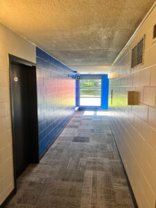 Vibrant hallway at Niagara College Canada’s Welland Campus, with blue brick walls, patterned carpeting, an emergency exit sign, and a window at the end, renovated by ICON Restoration.