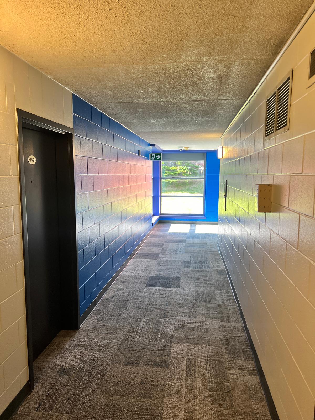 Vibrant hallway at Niagara College Canada’s Welland Campus, with blue brick walls, patterned carpeting, an emergency exit sign, and a window at the end, renovated by ICON Restoration.