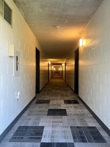 Modern hallway at Niagara College Canada’s Welland Campus, with new patterned carpeting, white brick walls, wall-mounted lights, and a smoke detector, post-renovation by ICON Restoration.
