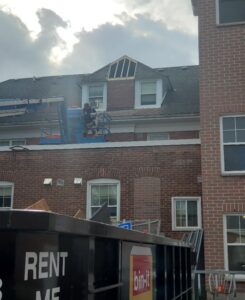 Image of a worker on a lift repairing the roof of a historic brick building, with a "Rent Me" bin-it dumpster in the foreground, under a partly cloudy sky.