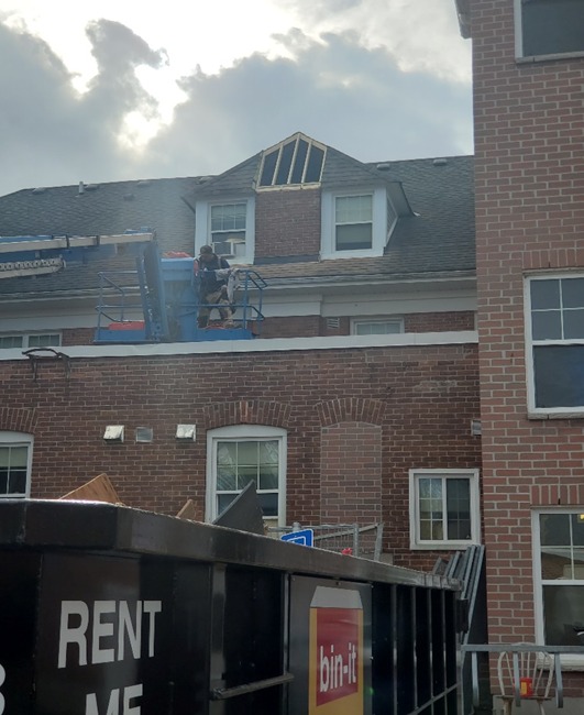 Image of a worker on a lift repairing the roof of a historic brick building, with a "Rent Me" bin-it dumpster in the foreground, under a partly cloudy sky.
