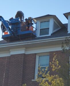 Image of workers on a lift conducting roof repair and maintenance on a historic brick building, with tools and safety equipment visible under a clear sky.