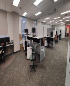 Image of an open office area with multiple adjustable desks, dual monitors, ergonomic chairs, and large windows, creating a collaborative workspace.