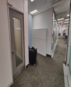 Image of a locker room with white lockers, a black chair with a cushion, and a frosted glass door, located in a modern office hallway.