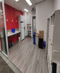 Image of a renovated break room with a red accent wall, white cabinets, a refrigerator, sink, and light wood flooring, featuring storage and cleaning supplies.