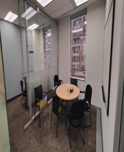 Image of a modern meeting room with glass walls, a round table, and several chairs, offering a view of the urban exterior through large windows.