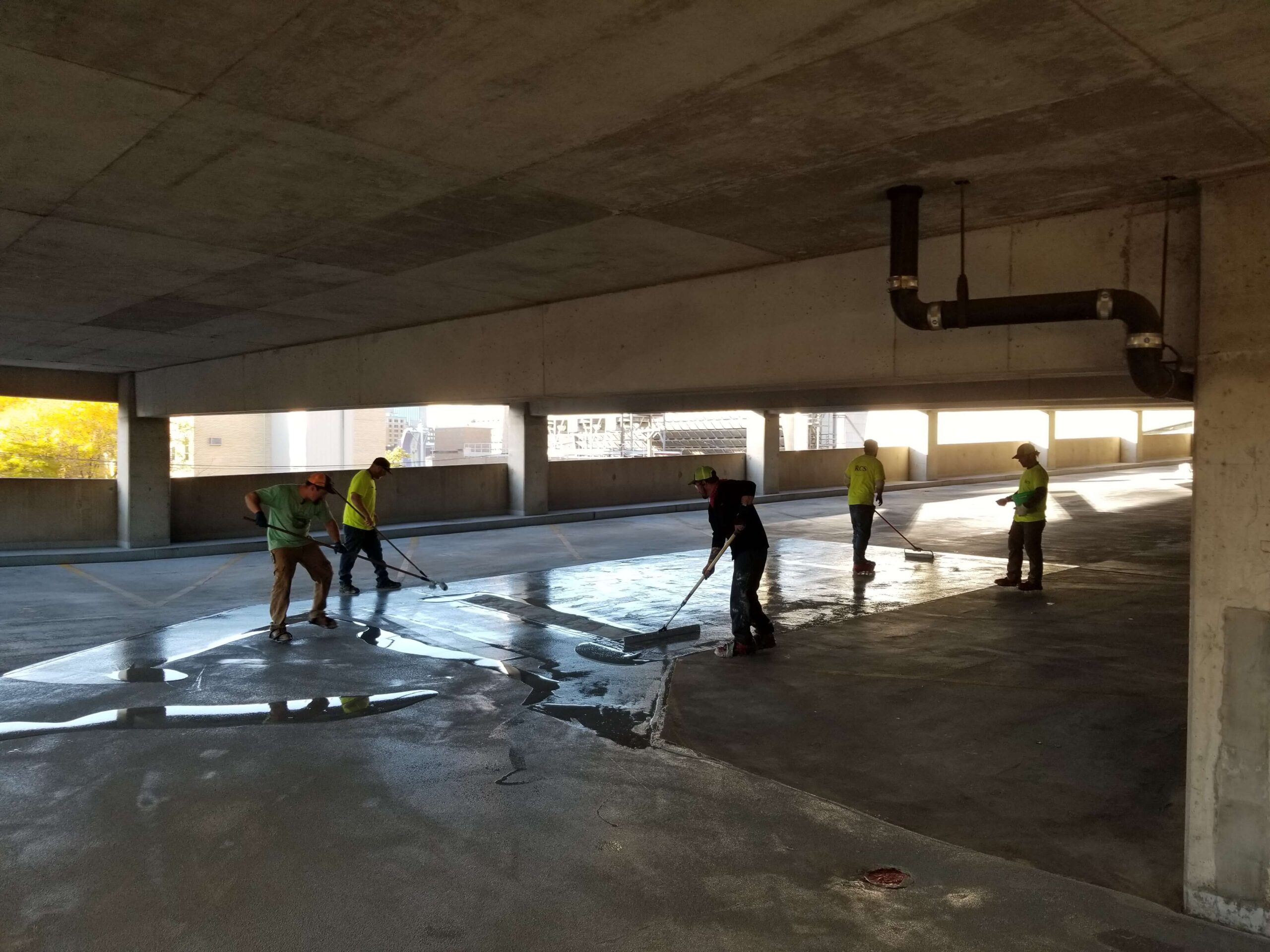 Workers in safety vests applying sealant to the floor of a parking garage with brooms.