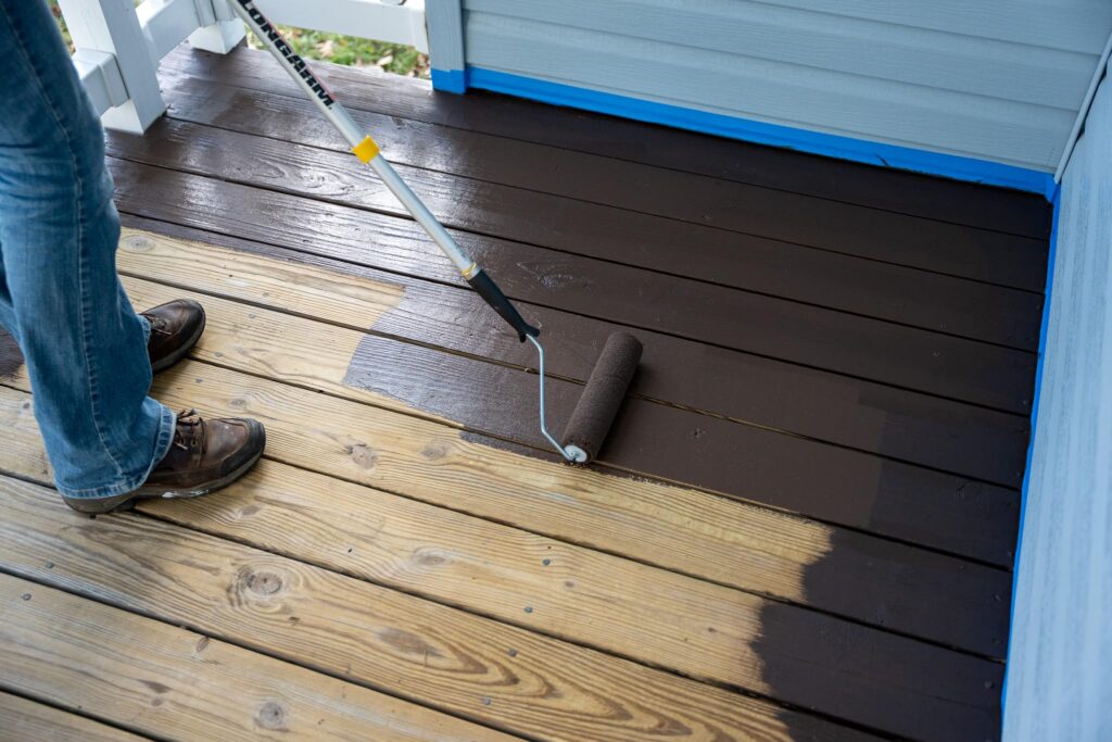 A person painting a wooden deck with a roller, applying dark stain to the wood surface.
