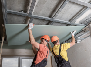 Two workers in safety gear installing drywall on a ceiling frame, wearing orange and yellow helmets and gloves.