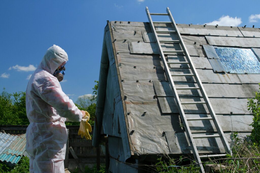 A person in a white hazmat suit with a respirator inspecting a weathered roof with a ladder under a blue sky.