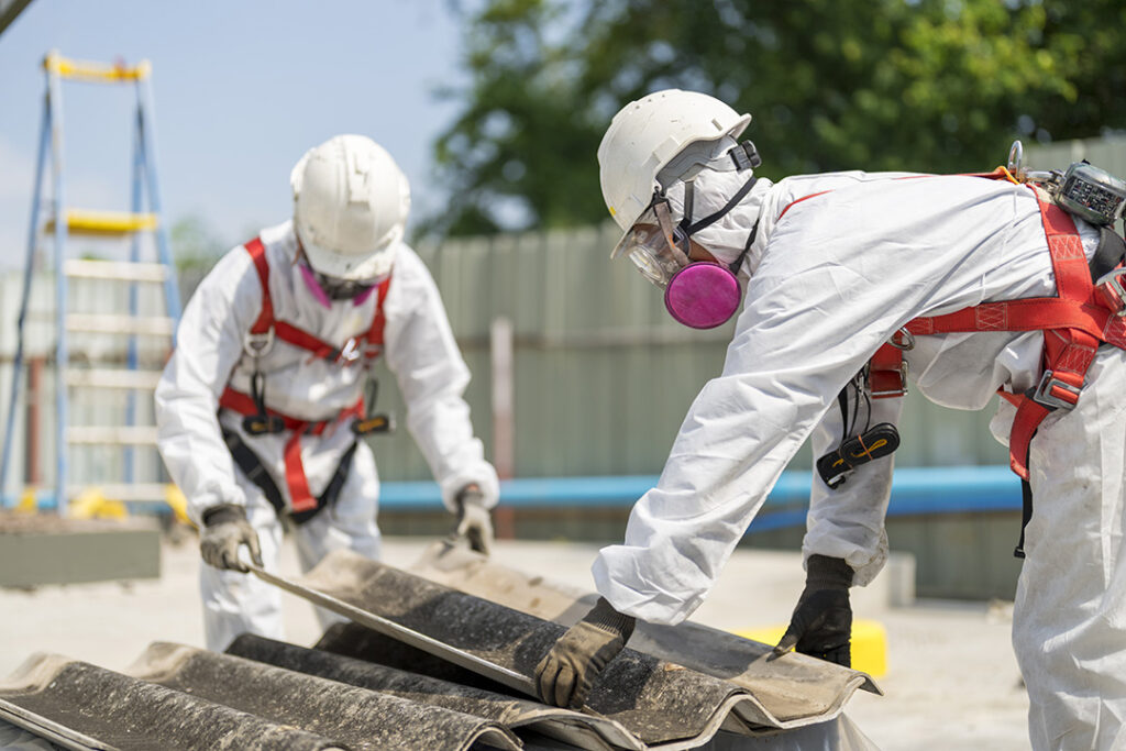 Two workers in white protective suits and helmets removing asbestos roof tiles with safety harnesses.