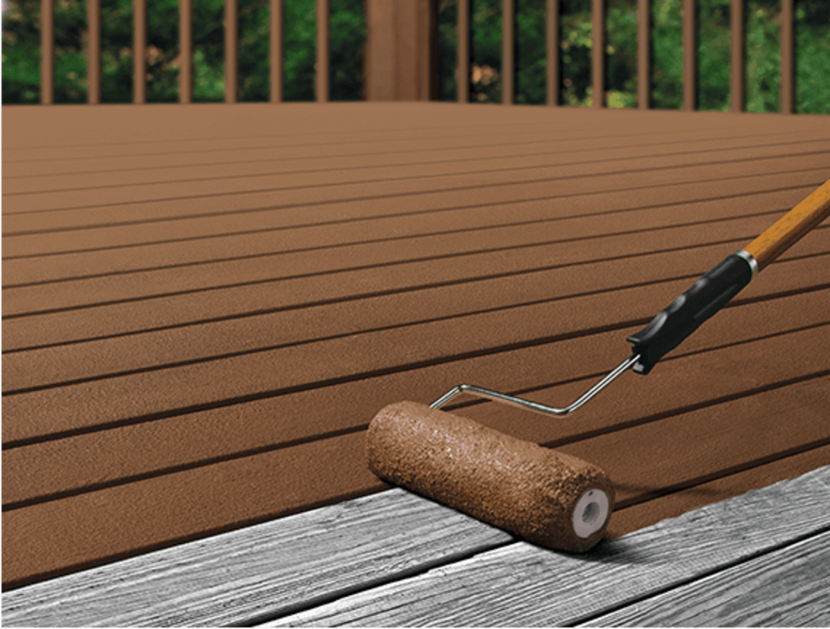 A roller applying brown sealant to a wooden deck with a paint can and bucket nearby.