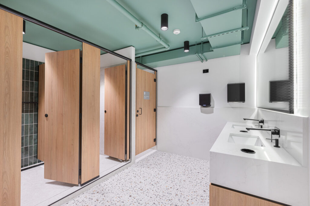 A clean, modern restroom with wooden stall doors, white sinks, and a green ceiling with recessed lighting.