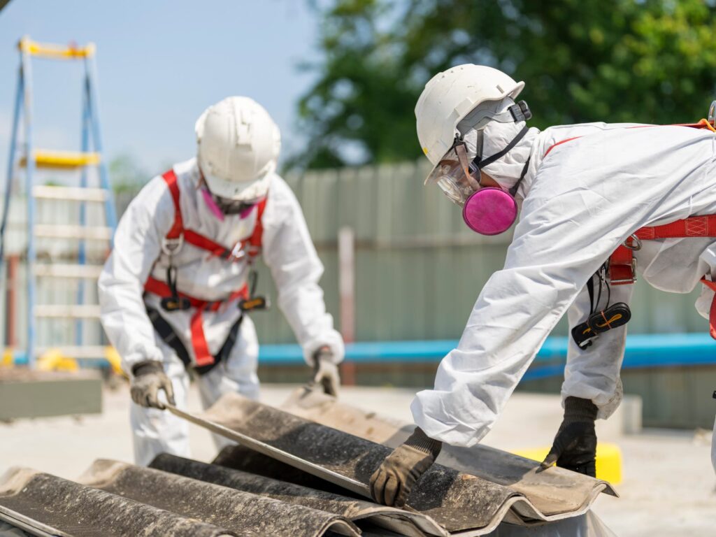 Two workers in white hazmat suits and helmets removing asbestos sheets with safety harnesses near a ladder.