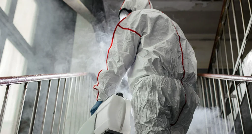 A worker in a white protective suit with red trim using a fogging machine to disinfect a stairwell filled with mist.
