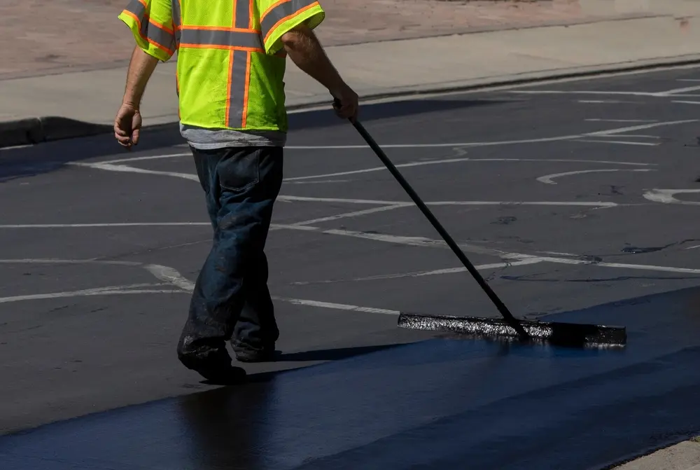A worker in a high-visibility vest smoothing fresh asphalt on a road with a long-handled tool.